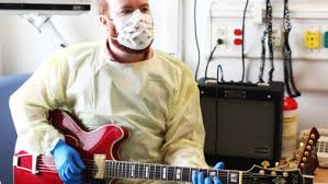 A man wearing a mask and hospital gown while holding a guitar